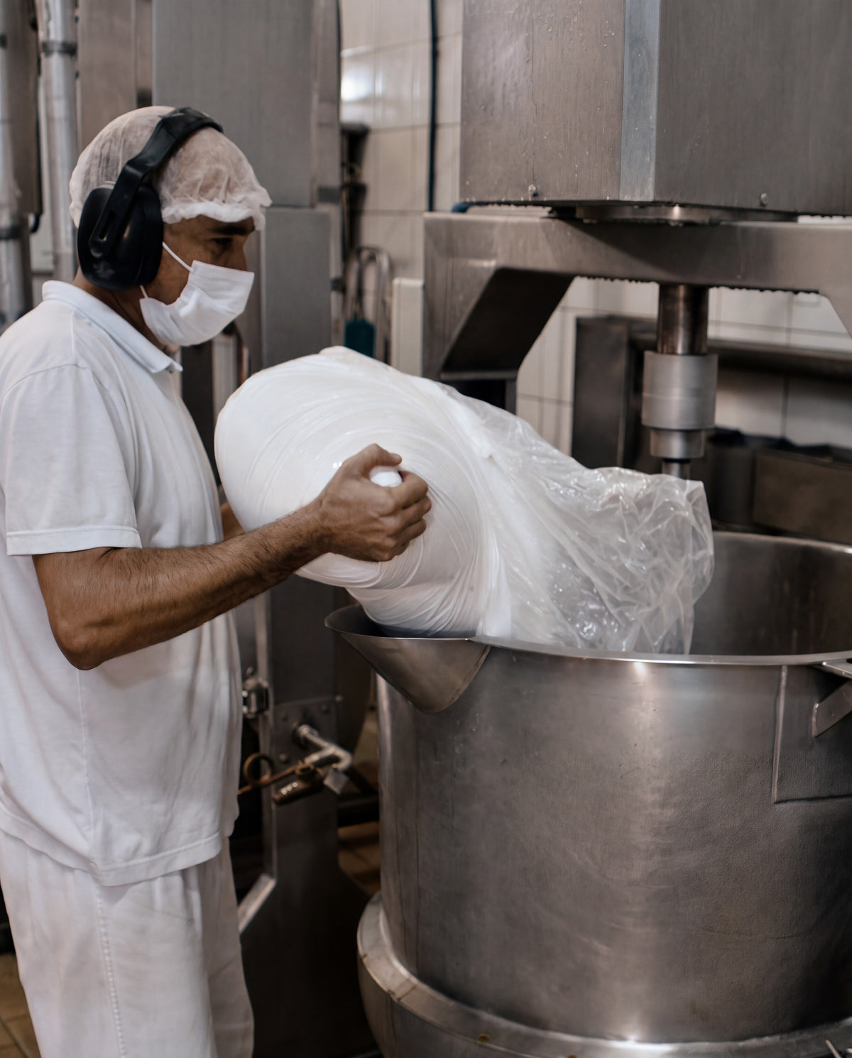 Operador de touca, máscara e protetor auricular despeja ingredientes no tacho de inox da produção de caramelos